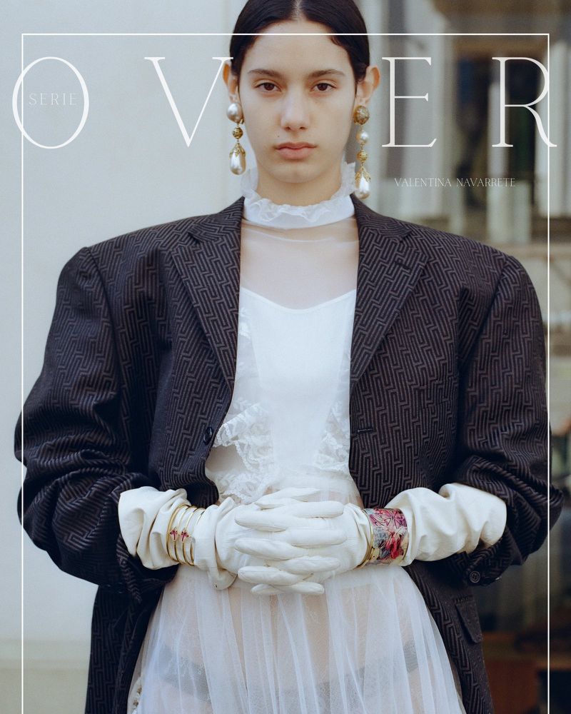 Editorial fashion shot: A person in an oversized blazer and white dress strikes a pose in front of a bright background and ornate building.