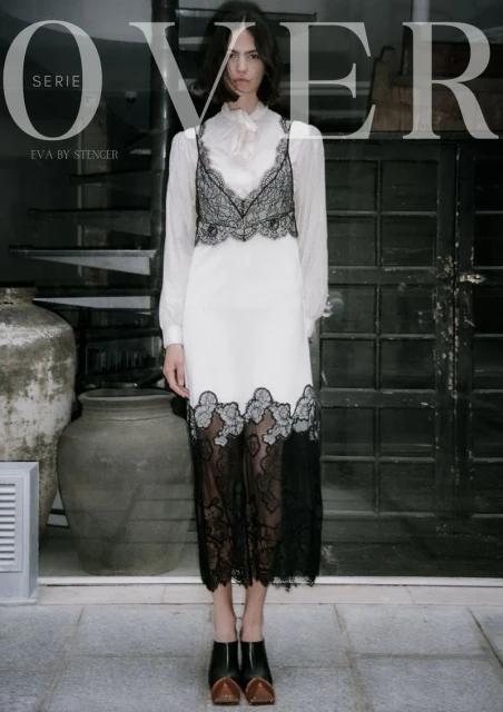 A female fashion model wearing a white dress with black lace accents, posing indoors with stone floors and textured walls, looking at the camera.