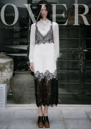 A female fashion model wearing a white dress with black lace accents, posing indoors with stone floors and textured walls, looking at the camera.