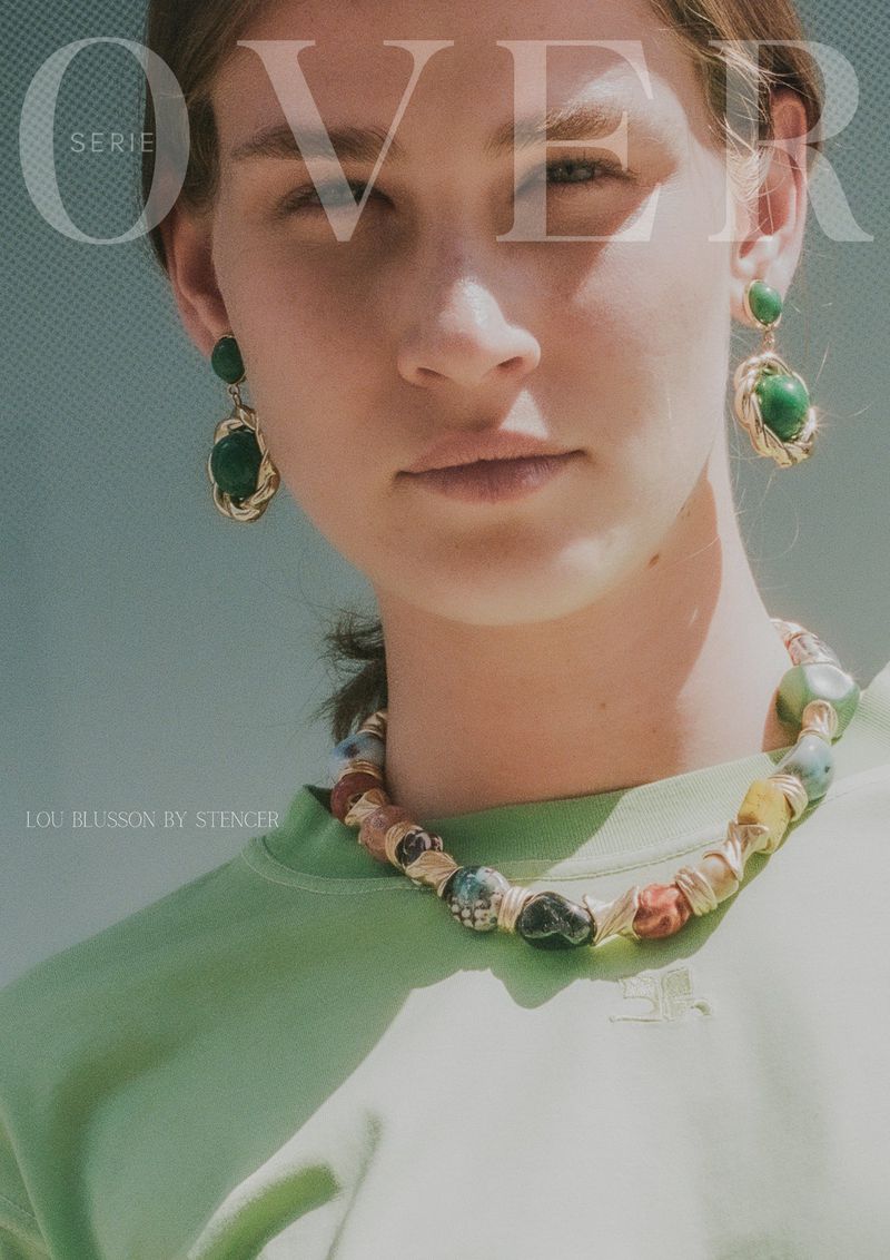Close-up of a woman with green earrings and necklace, wearing a light green shirt against a light blue background.