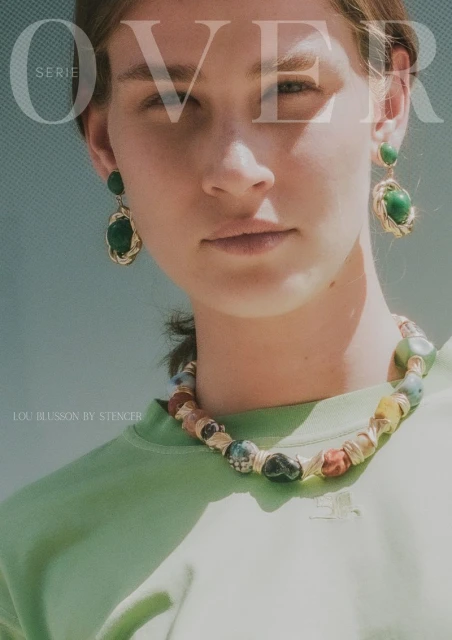 Close-up of a woman with green earrings and necklace, wearing a light green shirt against a light blue background.