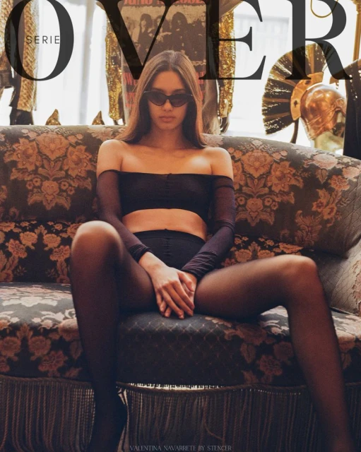 Portrait of a slender woman with dark hair wearing sunglasses and black lingerie, posing on a floral couch in an indoor setting.