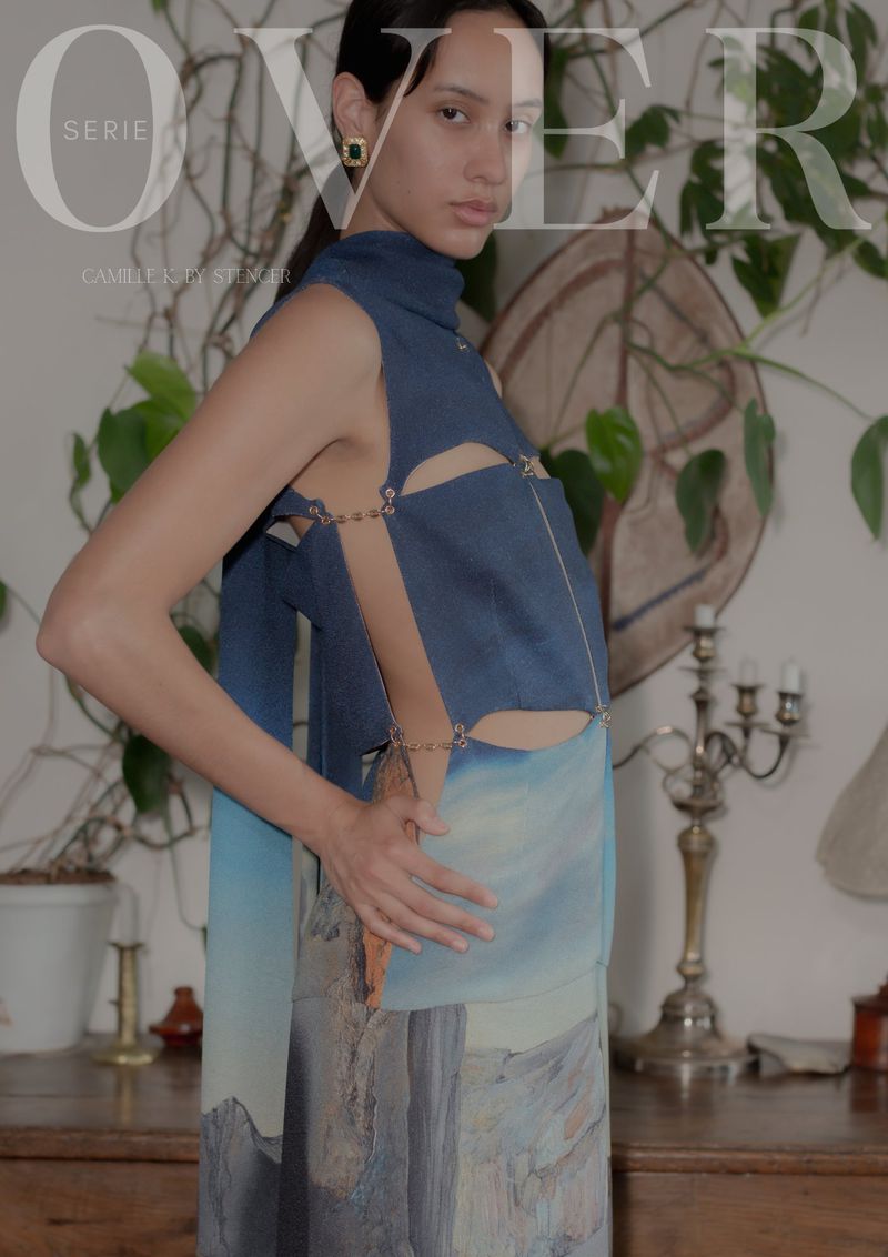 Elegant young woman poses indoors wearing a blue halter top and blue, white, and brown skirt in front of a plant and table.
