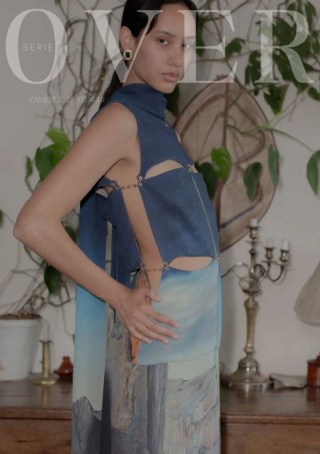 Elegant young woman poses indoors wearing a blue halter top and blue, white, and brown skirt in front of a plant and table.