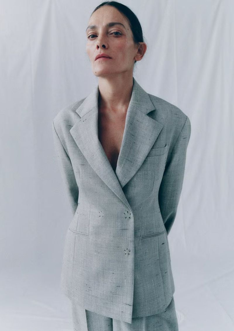 Woman in a grey double-breasted blazer and matching pants poses against a white backdrop in a studio setting, fashion portrait.