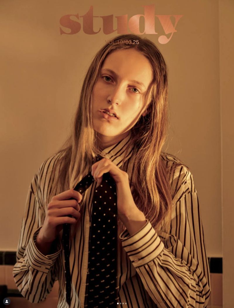 Portrait of a blonde-haired woman in a striped shirt adjusting a black tie in an indoor setting with warm lighting.