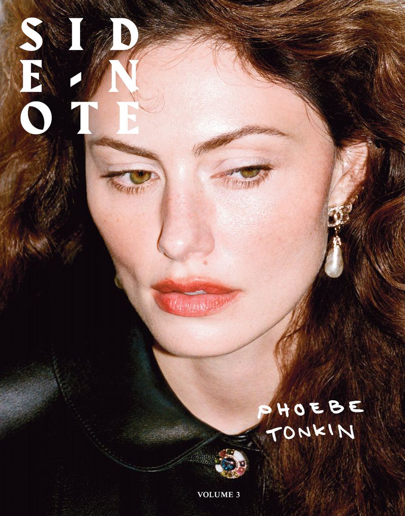 Close-up portrait of Phoebe Tonkin, with brown wavy hair, wearing a black leather top and gold earrings against a neutral backdrop.