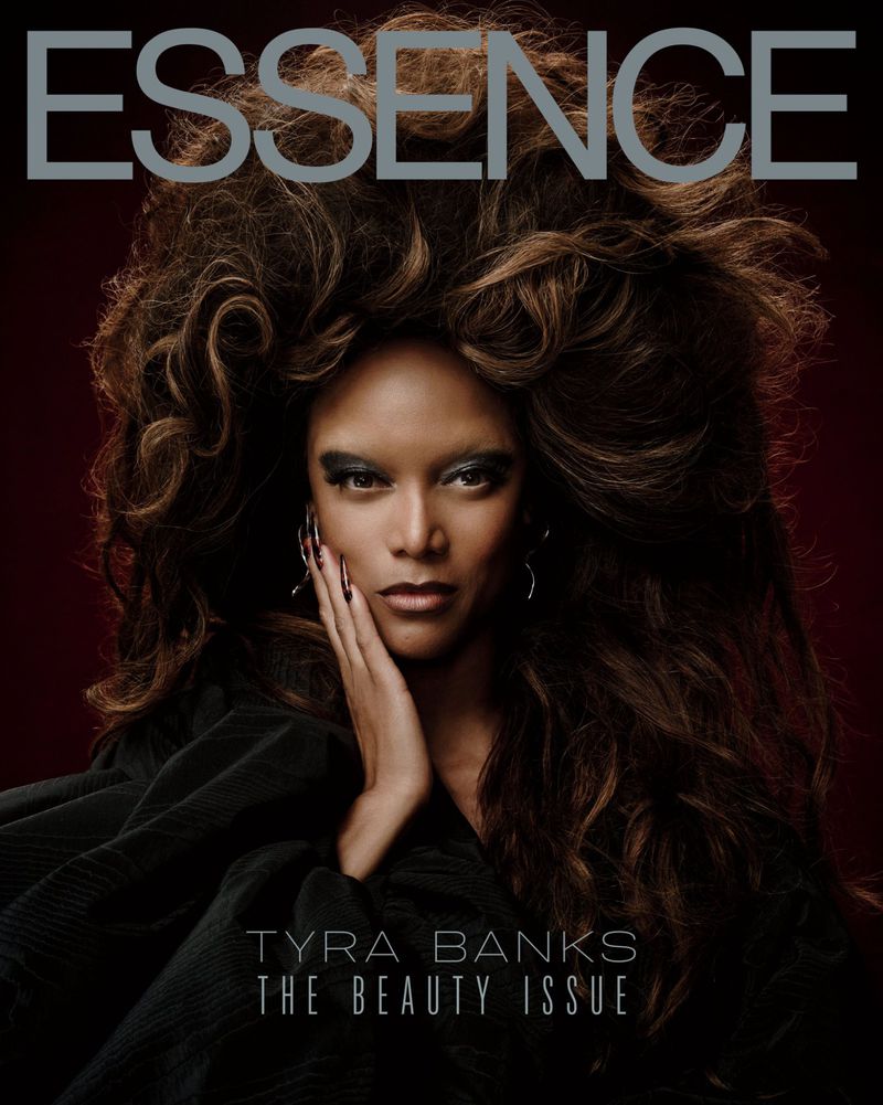 Essence magazine cover featuring Tyra Banks with voluminous brown hair and a dark outfit, against a deep red backdrop.