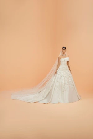 A woman wearing a white wedding dress with a long train and veil is posing in a studio with a peach background. Bridal fashion.