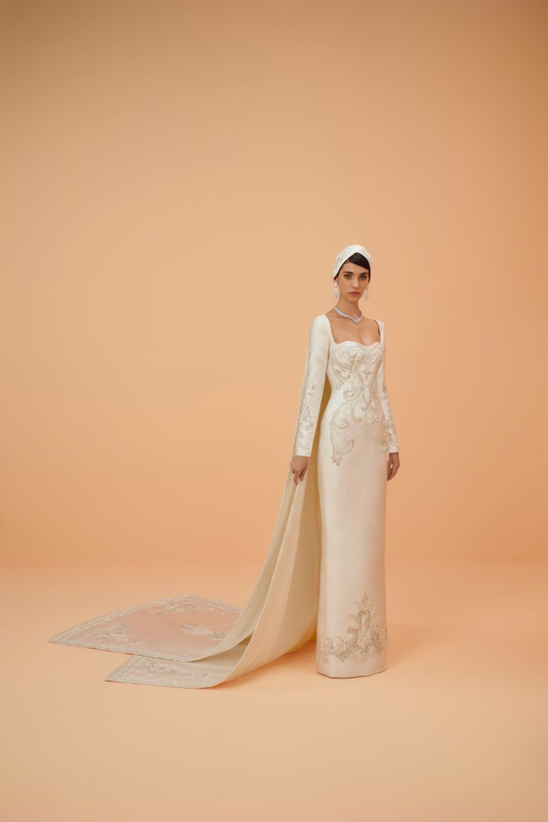 Elegant adult woman wearing an embroidered, long-sleeved, white couture gown with a head covering poses in a bright photo studio.