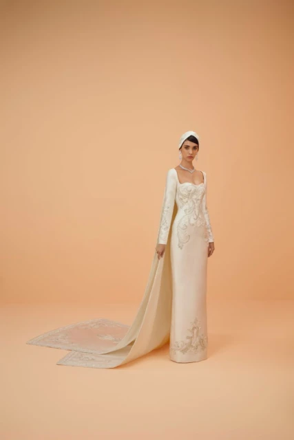 Elegant adult woman wearing an embroidered, long-sleeved, white couture gown with a head covering poses in a bright photo studio.