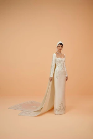 Elegant adult woman wearing an embroidered, long-sleeved, white couture gown with a head covering poses in a bright photo studio.