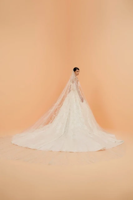 Elegant woman in white lace wedding gown and long veil poses in a bright studio with soft lighting and peach-toned backdrop. 