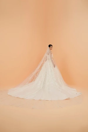Elegant woman in white lace wedding gown and long veil poses in a bright studio with soft lighting and peach-toned backdrop. 