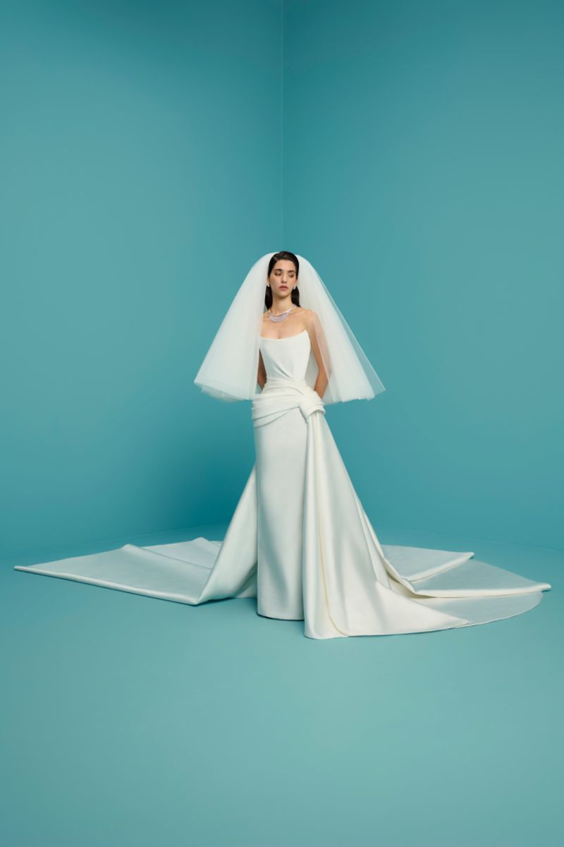 Woman in a white strapless gown and tulle veil poses in a studio against a blue background. Bridal fashion editorial with a contemporary look.