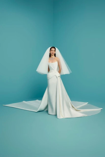 Woman in a white strapless gown and tulle veil poses in a studio against a blue background. Bridal fashion editorial with a contemporary look.