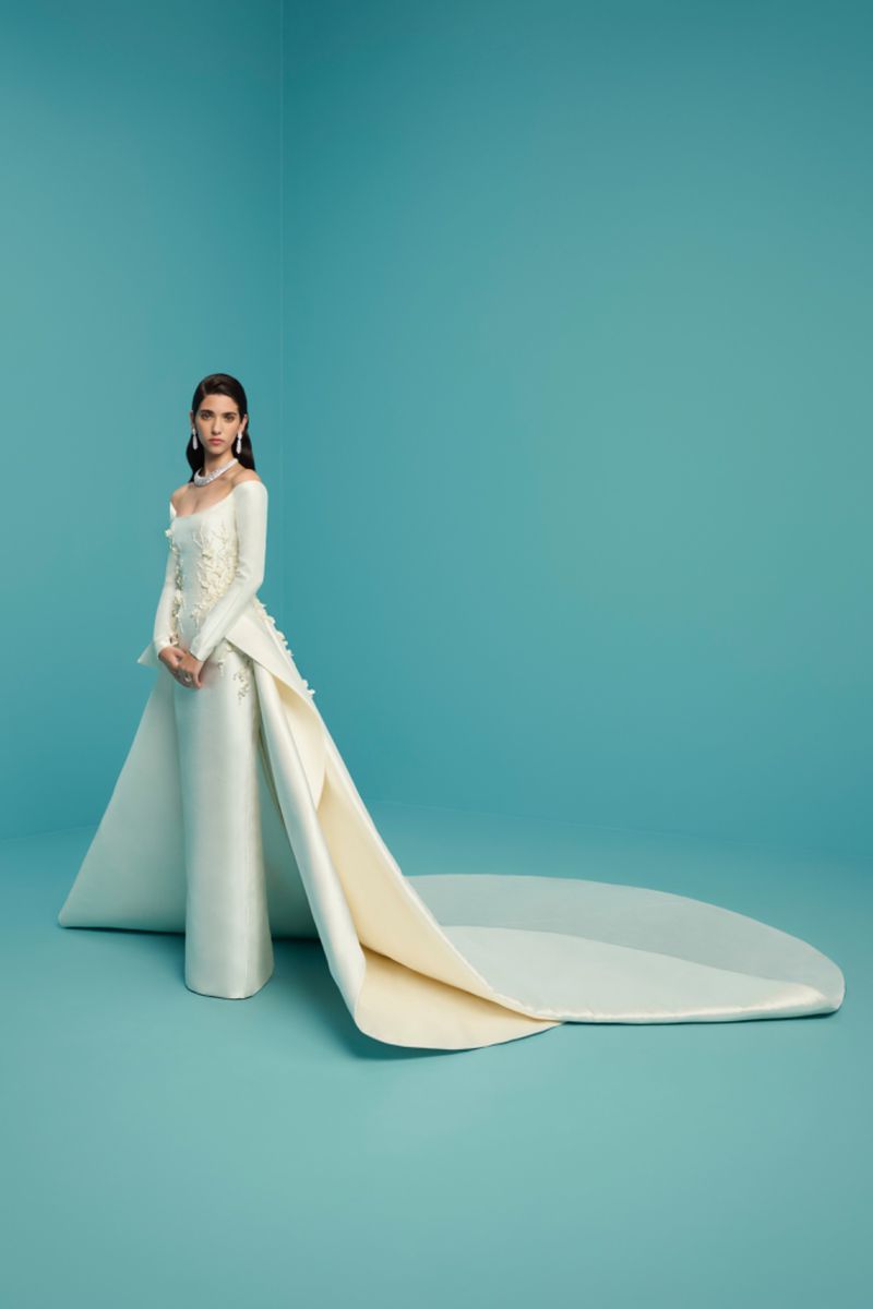Elegant woman in long-sleeved ivory gown with train, posing against a turquoise backdrop in a studio. Fashion photography.