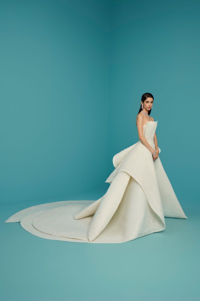 A woman stands in a formal cream-colored dress against a blue background, featuring a flowing train and elegant, simple styling.