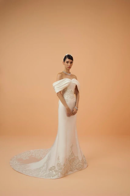A bride poses in a white off-the-shoulder wedding gown with a train on an orange background in a studio setting, looking at the camera.