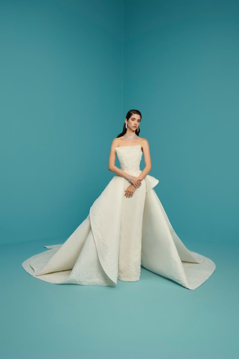 Model in a structured white strapless wedding gown with an extended overskirt standing against a teal studio backdrop.