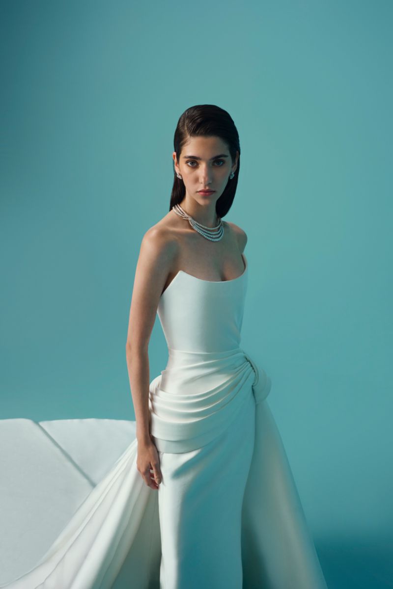 Portrait of a young adult woman wearing a white strapless dress and a layered pearl necklace, posing in front of a light blue backdrop.
