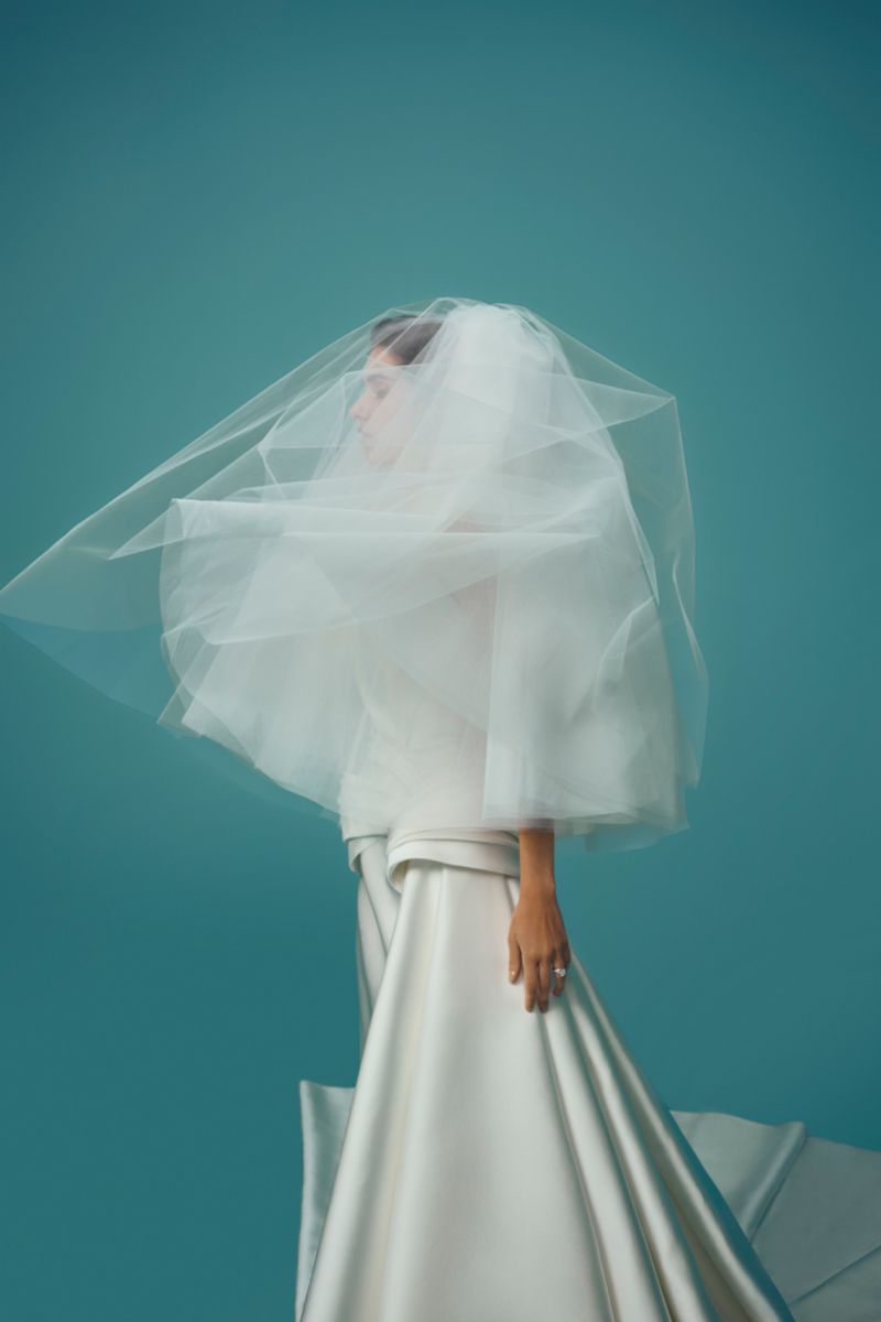 Model in white wedding dress and flowing veil poses in a studio with teal backdrop, fashion editorial, beauty shot, elegant and modern.