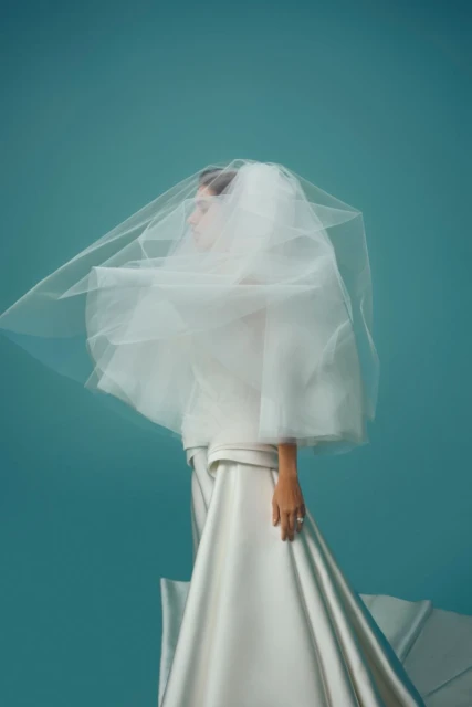 Model in white wedding dress and flowing veil poses in a studio with teal backdrop, fashion editorial, beauty shot, elegant and modern.