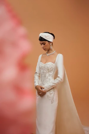 Elegant woman poses in long-sleeved white wedding gown with train and embellished bodice, wearing matching headpiece, against peach background.