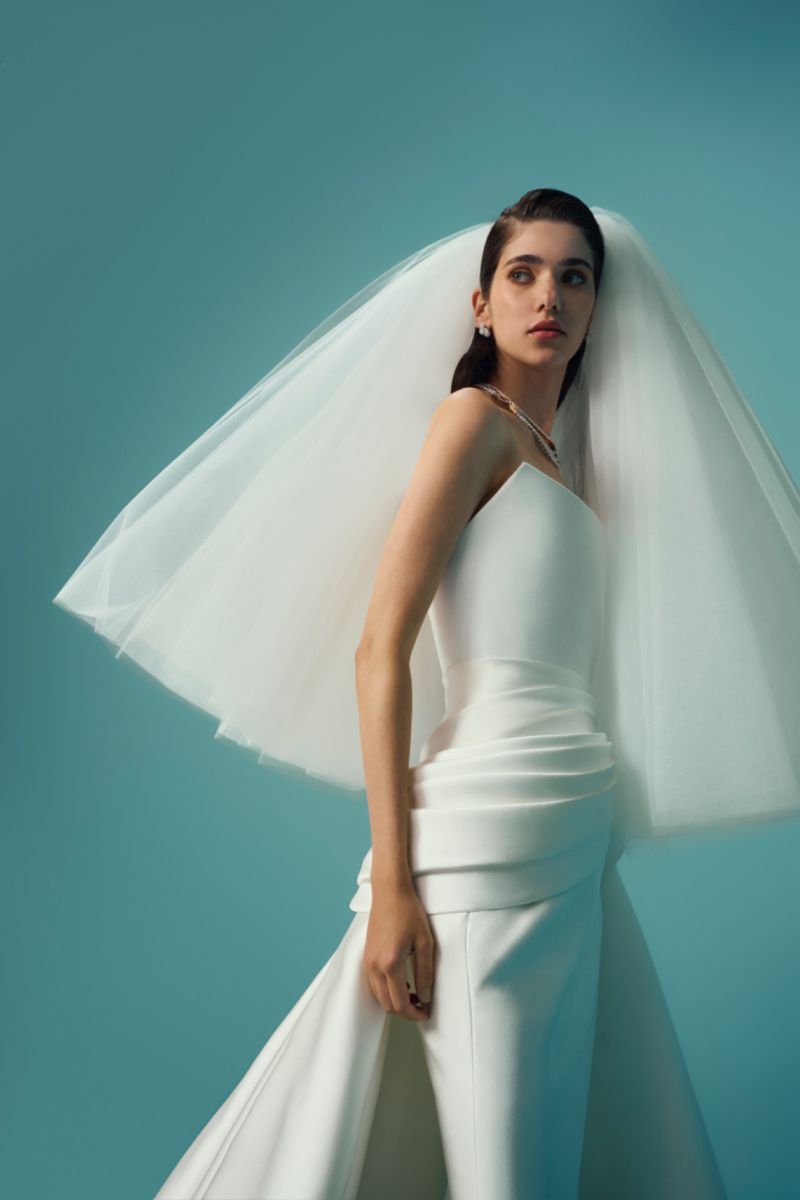 A stylish adult female bride poses in a white dress and veil, set against a bright blue background, emphasizing fashion and elegance.