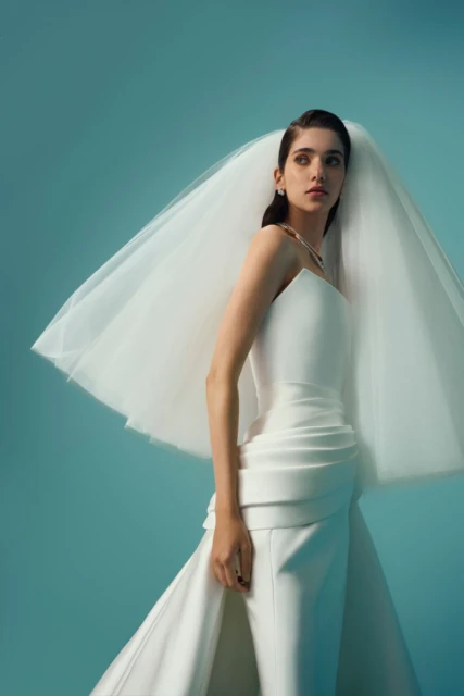 A stylish adult female bride poses in a white dress and veil, set against a bright blue background, emphasizing fashion and elegance.