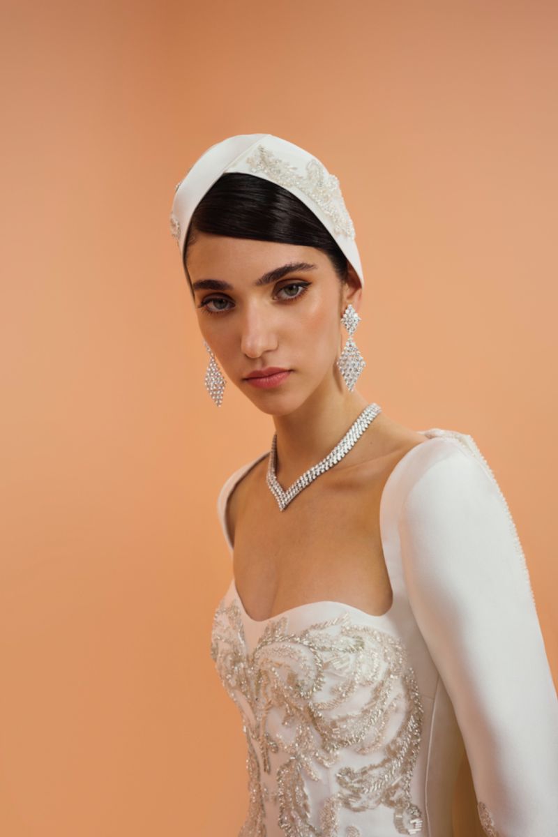 Elegant woman in a white headscarf, gown, and jewelry posing against a peach background in a studio portrait.