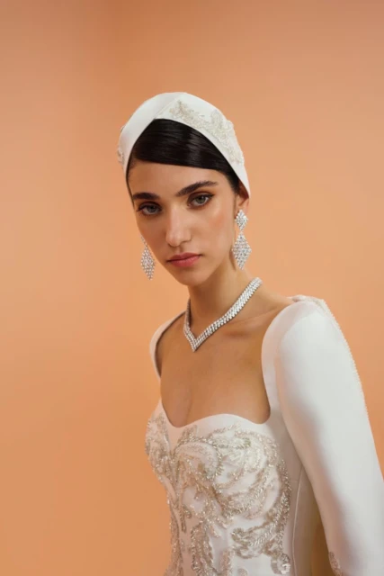 Elegant woman in a white headscarf, gown, and jewelry posing against a peach background in a studio portrait.