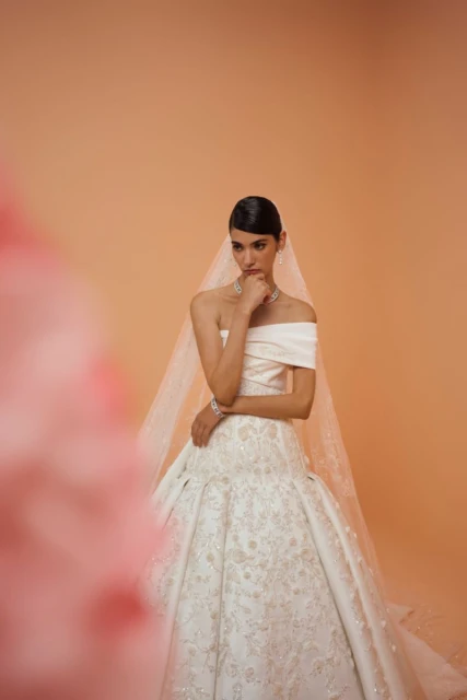 Elegant woman in an off-the-shoulder white wedding gown with veil posing against a peach background and pink floral foreground.