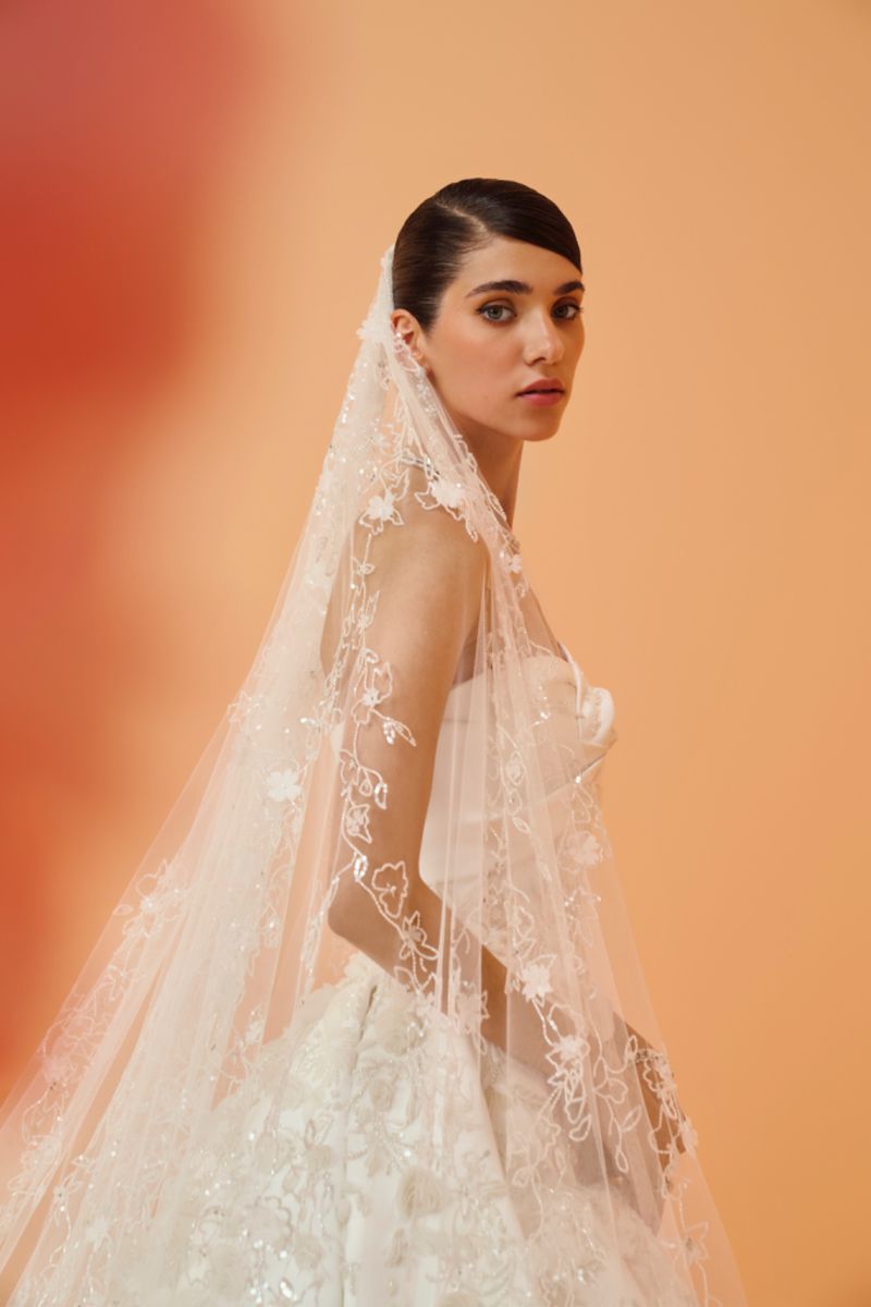 Elegant woman in strapless wedding gown and floral veil posing in a portrait against a peach colored background in a studio.