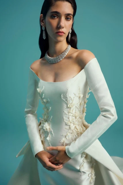 A brunette woman in a white gown and diamond jewelry poses in a studio with a teal background. The gown has long sleeves.