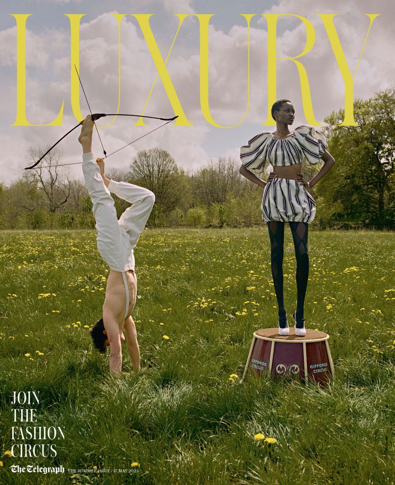 Fashion editorial image: a shirtless man balances upside down holding a bow and arrow, with an African-American model standing on a drum.