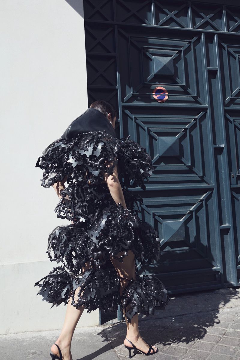 Model wearing a voluminous black sculptural ruffled dress and slingback kitten heels walking past a dark geometric doorway.