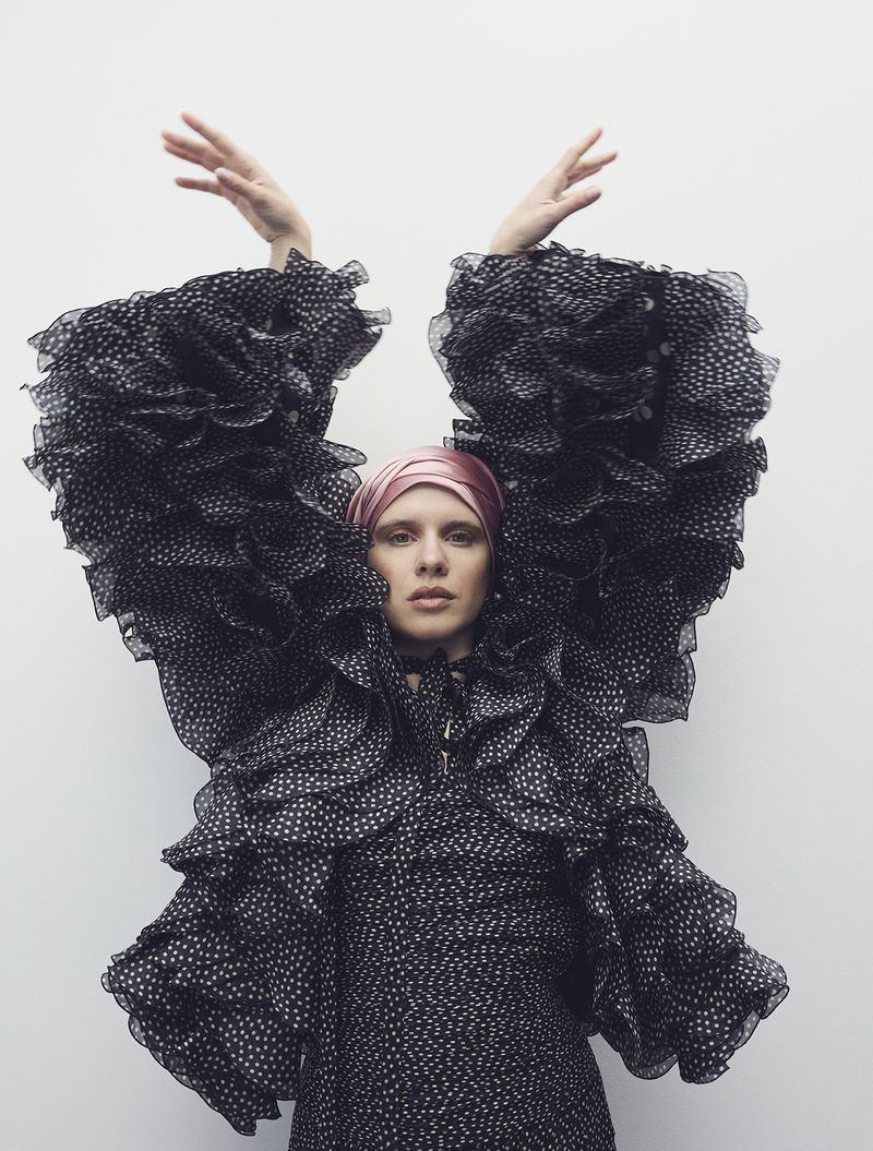 Woman in a polka dot dress with large ruffled sleeves, wearing a pink headscarf, standing against a white wall, raising her hands.