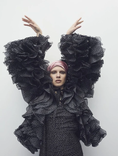 Woman in a polka dot dress with large ruffled sleeves, wearing a pink headscarf, standing against a white wall, raising her hands.