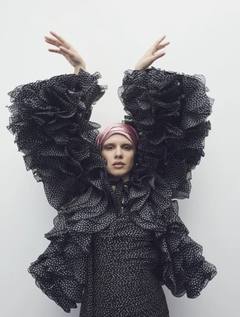 Woman in a polka dot dress with large ruffled sleeves, wearing a pink headscarf, standing against a white wall, raising her hands.