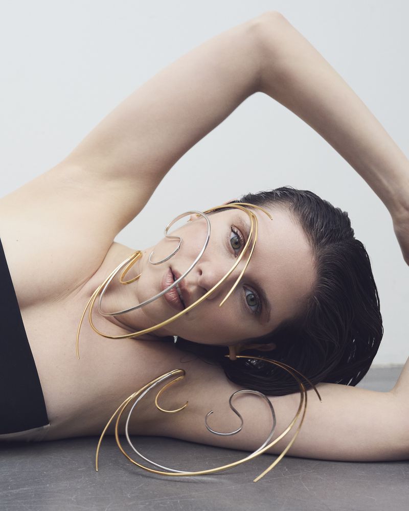Adult female model lies on a gray surface wearing artistic silver and gold metal jewelry. The model has dark hair and fair skin.