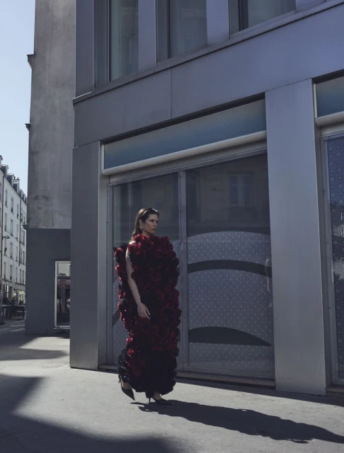 A woman in a red rose gown walking on a city sidewalk in daytime, showcasing a unique fashion statement with urban architecture.