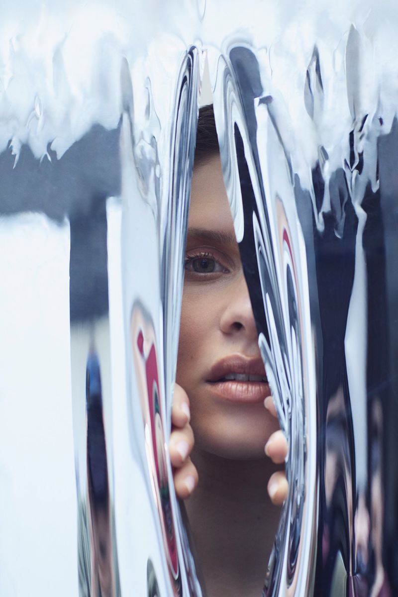 Close-up studio shot of an adult female peering through shimmering silver metallic curtains, creating distorted reflections on their surface.
