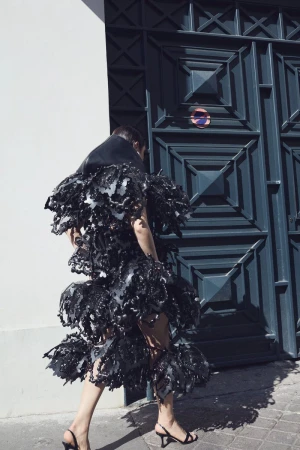 Model wearing a voluminous black sculptural ruffled dress and slingback kitten heels walking past a dark geometric doorway.
