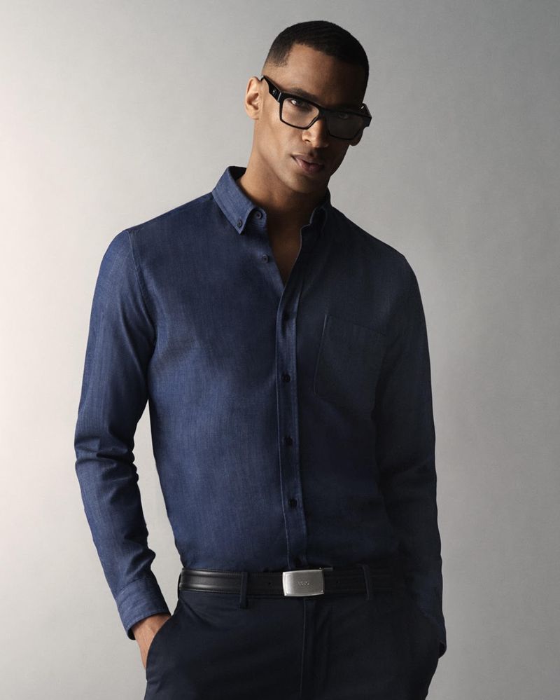 Studio shot of a dark-haired adult male wearing glasses, a long-sleeved denim shirt, and dark pants with a belt, posing against a neutral backdrop.
