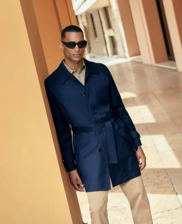 Dark-skinned adult male in sunglasses, blue trench coat, and tan pants leans against an orange column in an outdoor urban setting.
