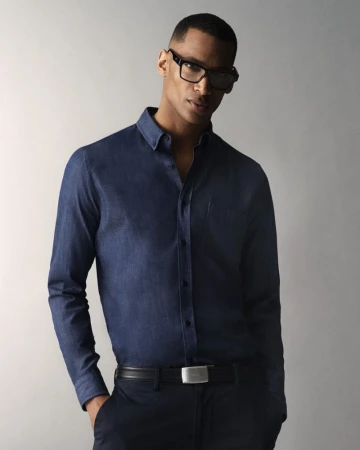 Man wearing a blue button-down shirt and black glasses standing against a neutral studio backdrop