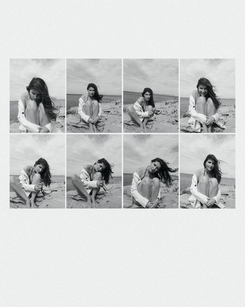 Black and white grid of a woman in a white jacket sitting on a sandy beach with rocks, with the sea and sky visible in the background.