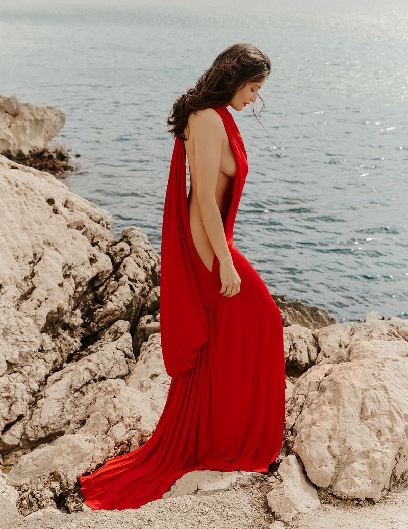 Laetitia Casta wearing a draped backless red gown while walking along a rocky Mediterranean coastline by the sea.