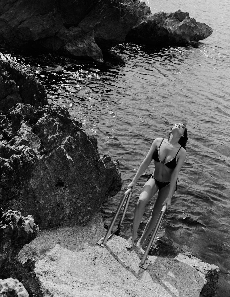 Black and white shot of a woman in a bikini climbing out of the ocean on a ladder, with rocks on the side, shot in natural light.